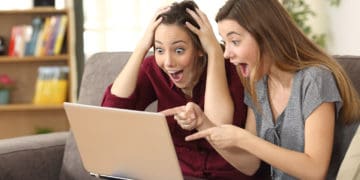 Two astonished roommates watching media content on line with a laptop sitting on a couch in the living room in a house interior