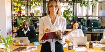 Photo by ANTONI SHKRABA from Pexels: https://www.pexels.com/photo/woman-standing-with-a-notebook-and-people-working-on-laptops-behind-her-in-an-office-7163456/